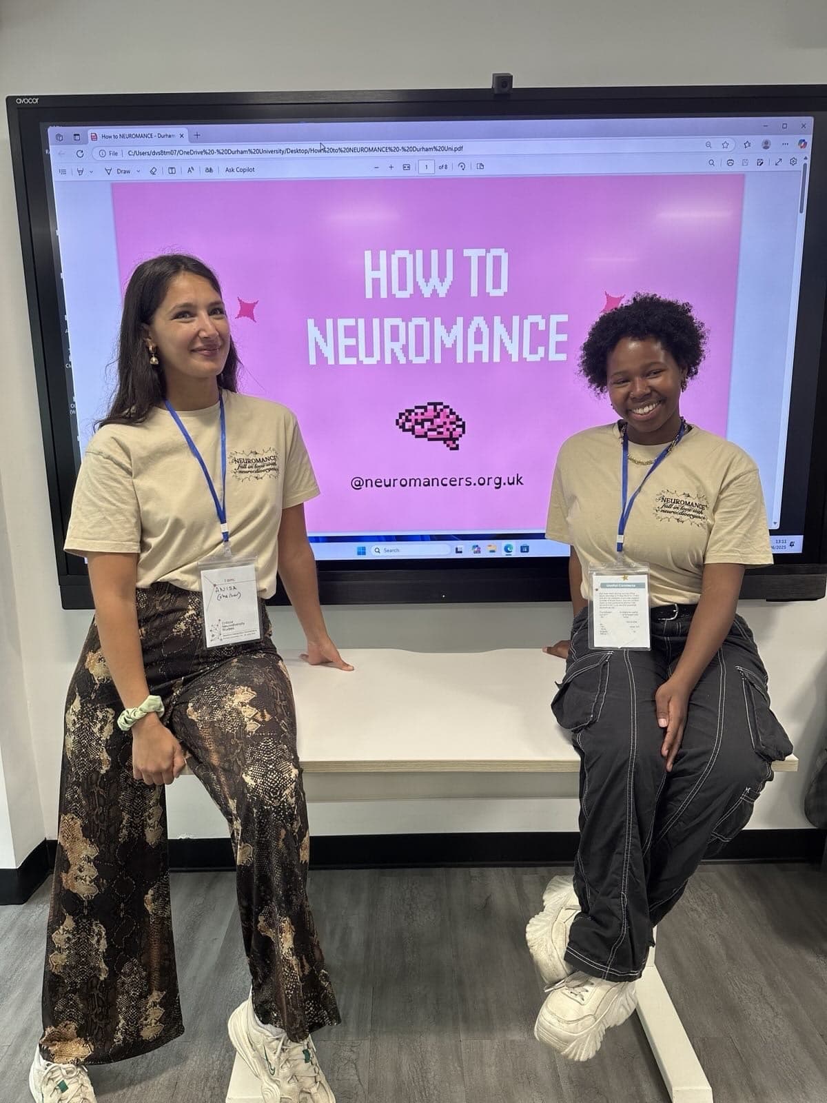 Two young individuals sit on a table with a screen behind them which says 'How To Neuromance'