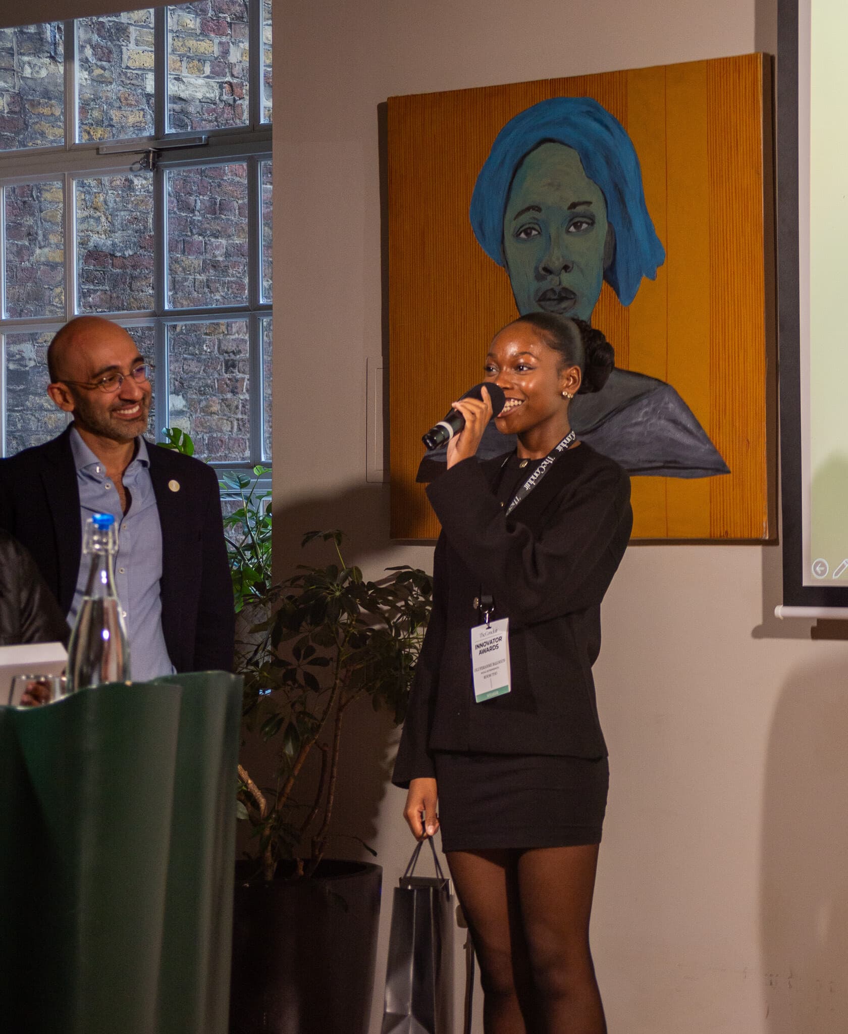 A female-presenting black individual speaks holding a microphone with a male-presenting individual looking on