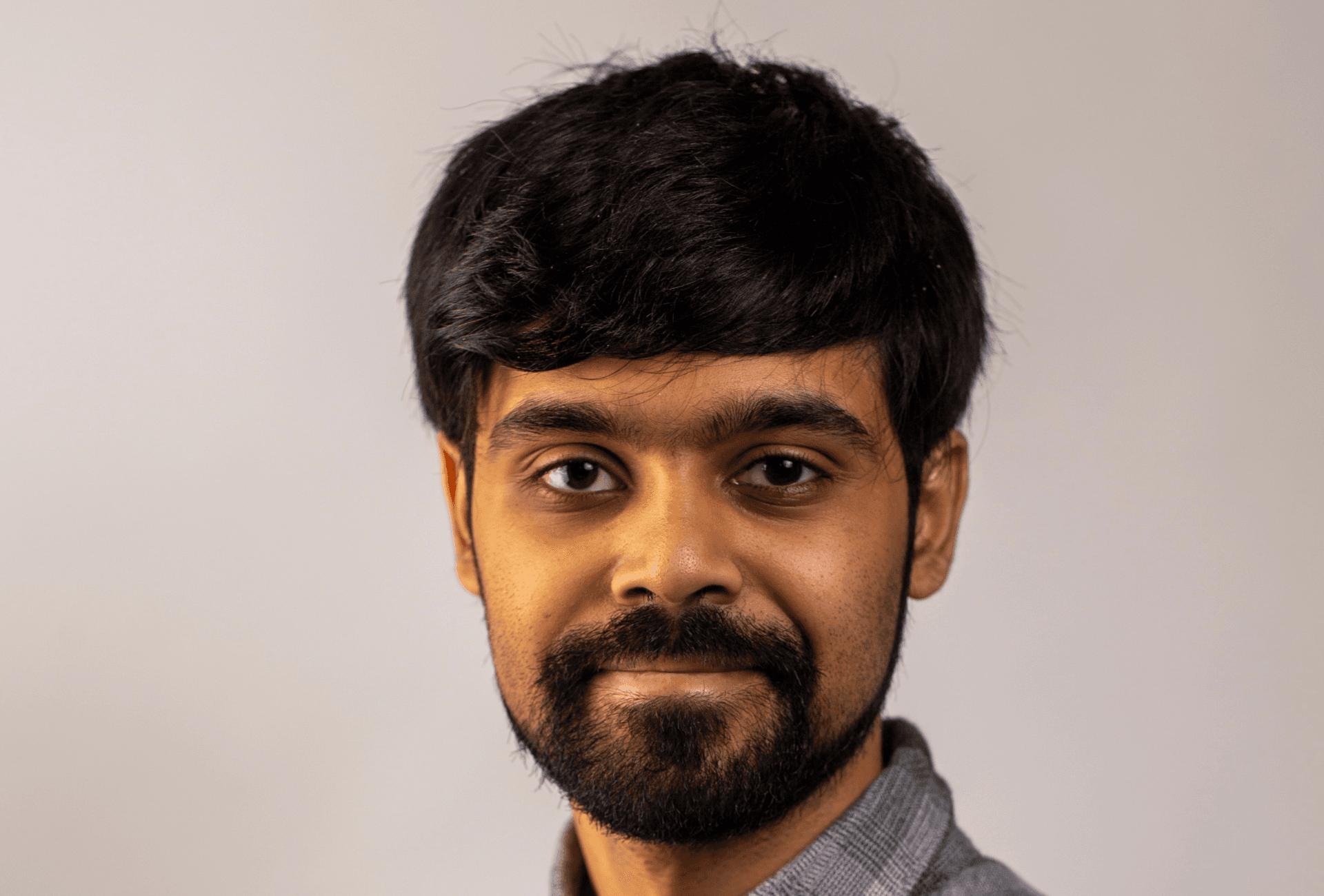 A headshot of a person with dark hair and a beard, smiling slightly at the camera. They are wearing a grey and black plaid button-down shirt, set against a neutral, light-grey background.